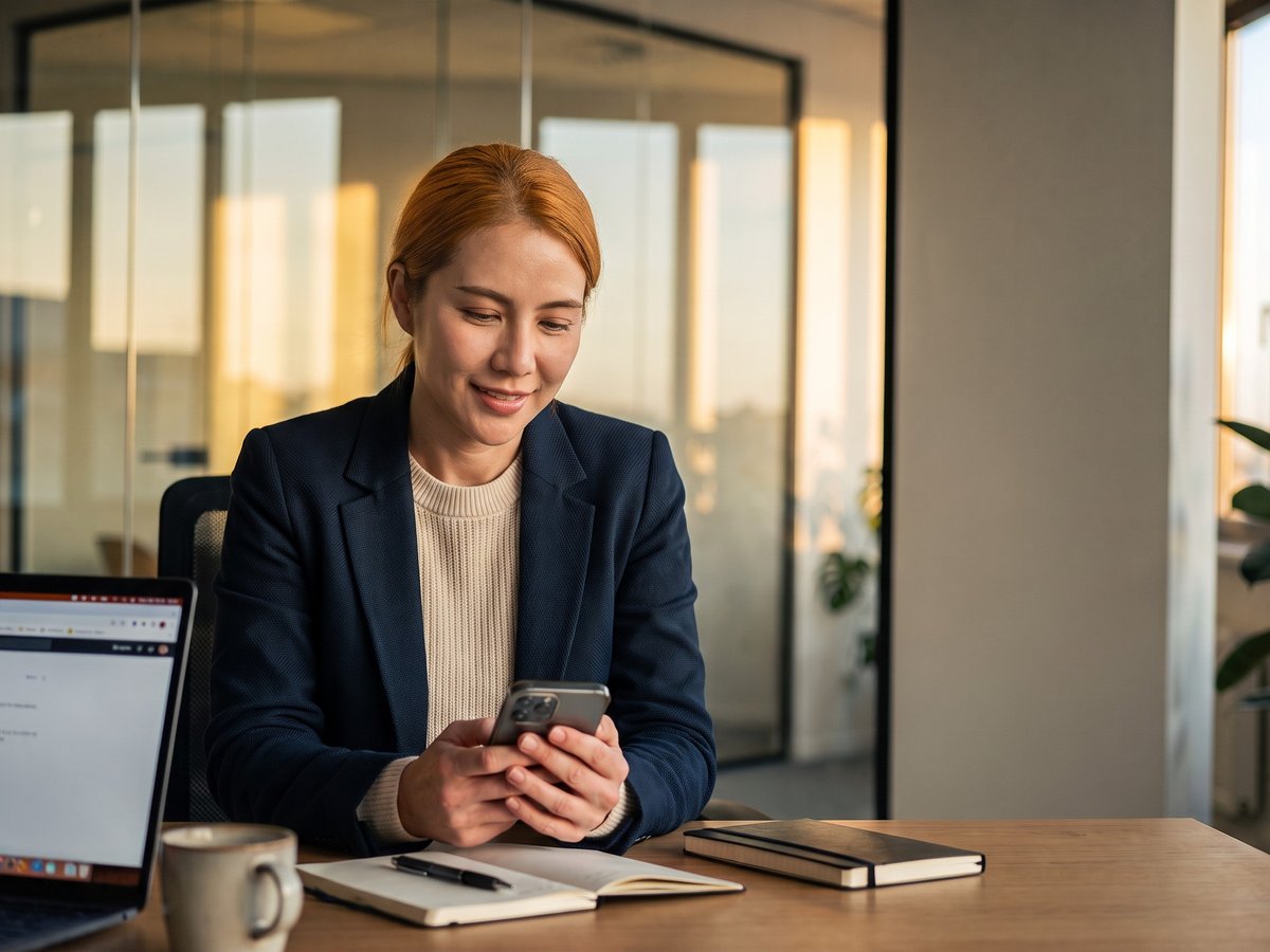 A professional smiling at her phone at her desk, with a laptop and notebook beside her